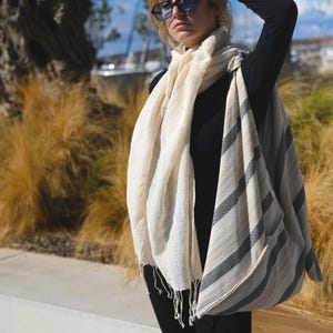 May include: A cream-colored scarf with gray and black stripes and fringe detail. The scarf is draped over a black long-sleeve top and black pants. The person is wearing sunglasses.