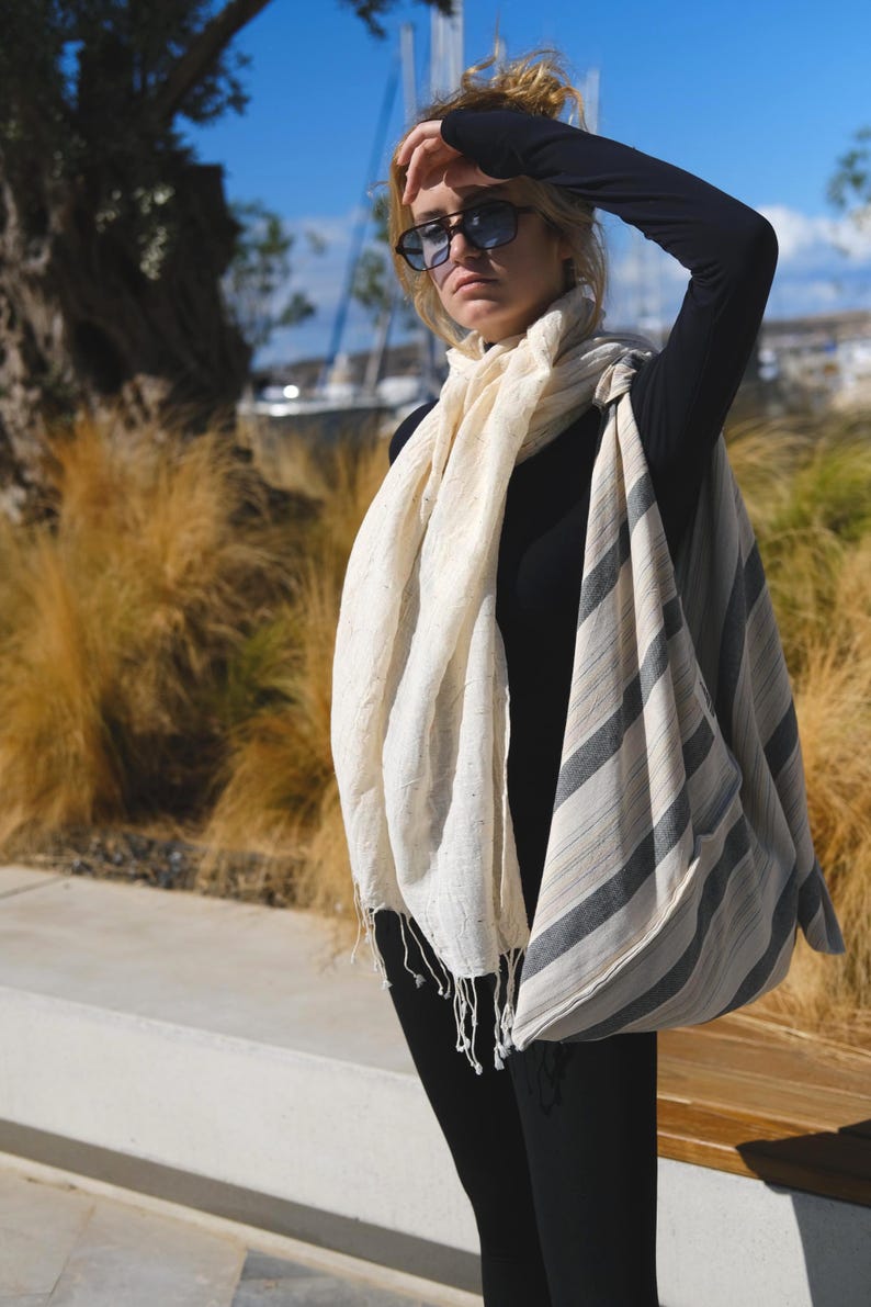 May include: A cream-colored scarf with gray and black stripes and fringe detail. The scarf is draped over a black long-sleeve top and black pants. The person is wearing sunglasses.