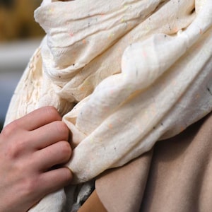 May include: A cream-colored scarf with flecks of orange, yellow, and black. The scarf is draped around the neck and held by a hand. The scarf appears to be made of a lightweight fabric.