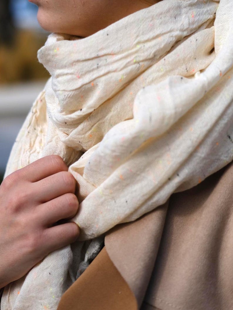 May include: A cream-colored scarf with flecks of orange, yellow, and black. The scarf is draped around the neck and held by a hand. The scarf appears to be made of a lightweight fabric.