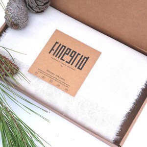 May include: A white linen and cotton scarf with fringed edges is presented in an open cardboard box. A brown tag with the text "FINEGRID" is placed on the scarf. Pine needles and pine cones are arranged around the box.