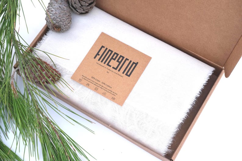 May include: A white linen and cotton scarf with fringed edges is presented in an open cardboard box. A brown tag with the text "FINEGRID" is placed on the scarf. Pine needles and pine cones are arranged around the box.