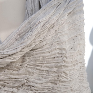 May include: A light gray scarf with a textured, crinkled appearance. The fabric has small, dark flecks throughout, adding visual interest. The scarf is draped, showcasing its soft, flowing nature. This accessory is suitable for various styles.