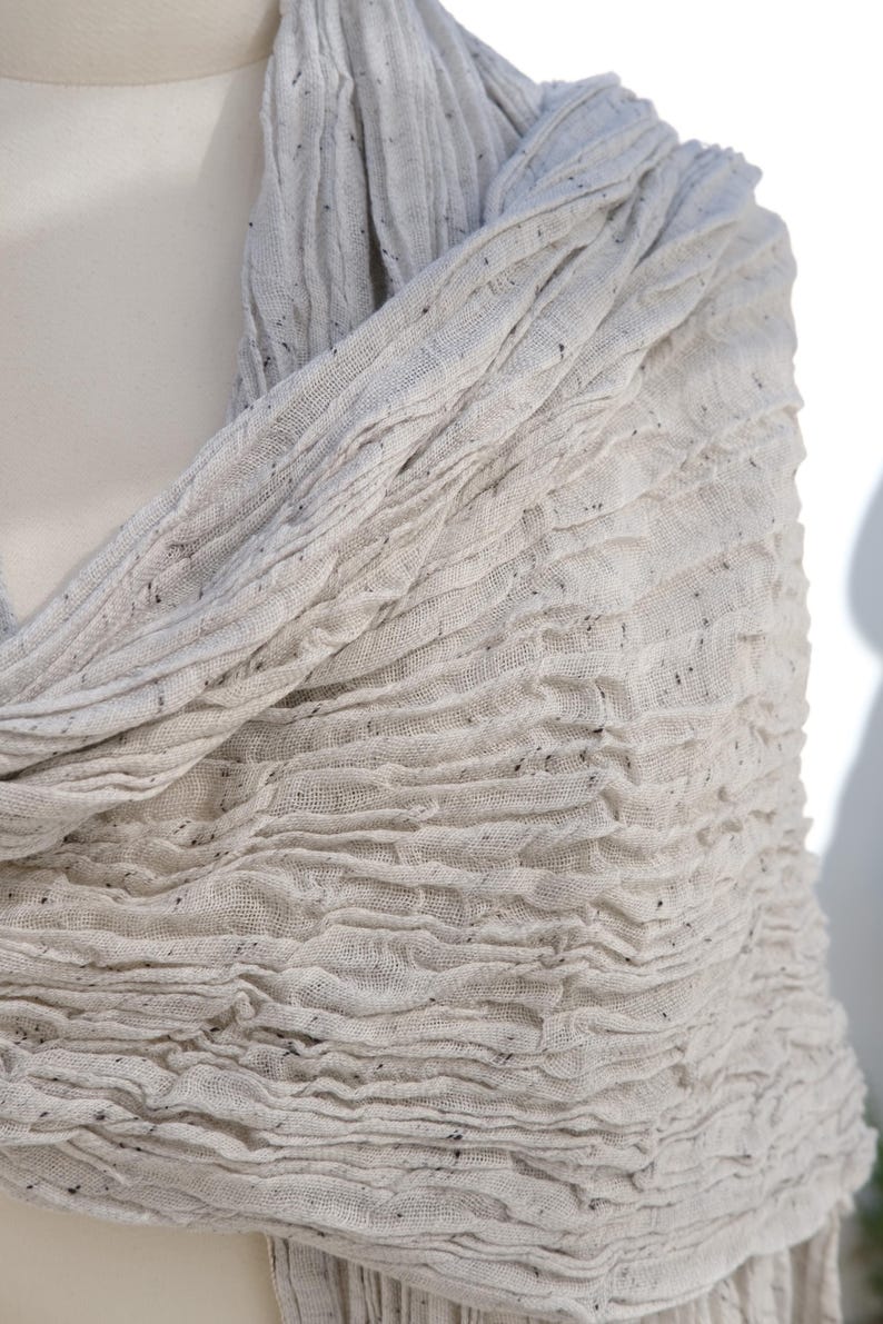 May include: A light gray scarf with a textured, crinkled appearance. The fabric has small, dark flecks throughout, adding visual interest. The scarf is draped, showcasing its soft, flowing nature. This accessory is suitable for various styles.