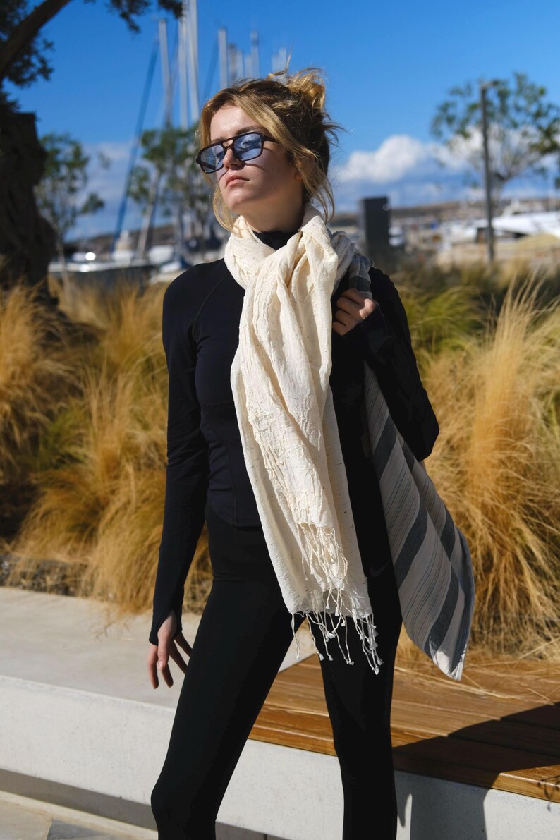 May include: A cream-colored scarf with fringe is draped over the shoulder. The scarf has a textured appearance and is paired with a black long-sleeve top and black leggings. The background features tall grass and a blue sky.