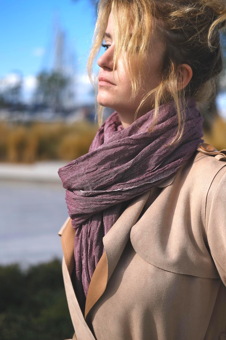 May include: A woman wearing a mauve scarf and a tan trench coat. The scarf is draped around the neck and shoulders. The coat has a classic design with a notched collar and a double-breasted closure.