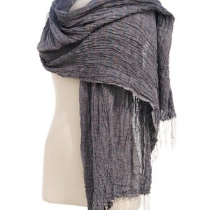 May include: A draped, textured scarf in shades of gray and blue, displayed on a mannequin. The scarf has a crinkled appearance and is finished with white fringe at the ends. The fabric appears lightweight and suitable for layering.