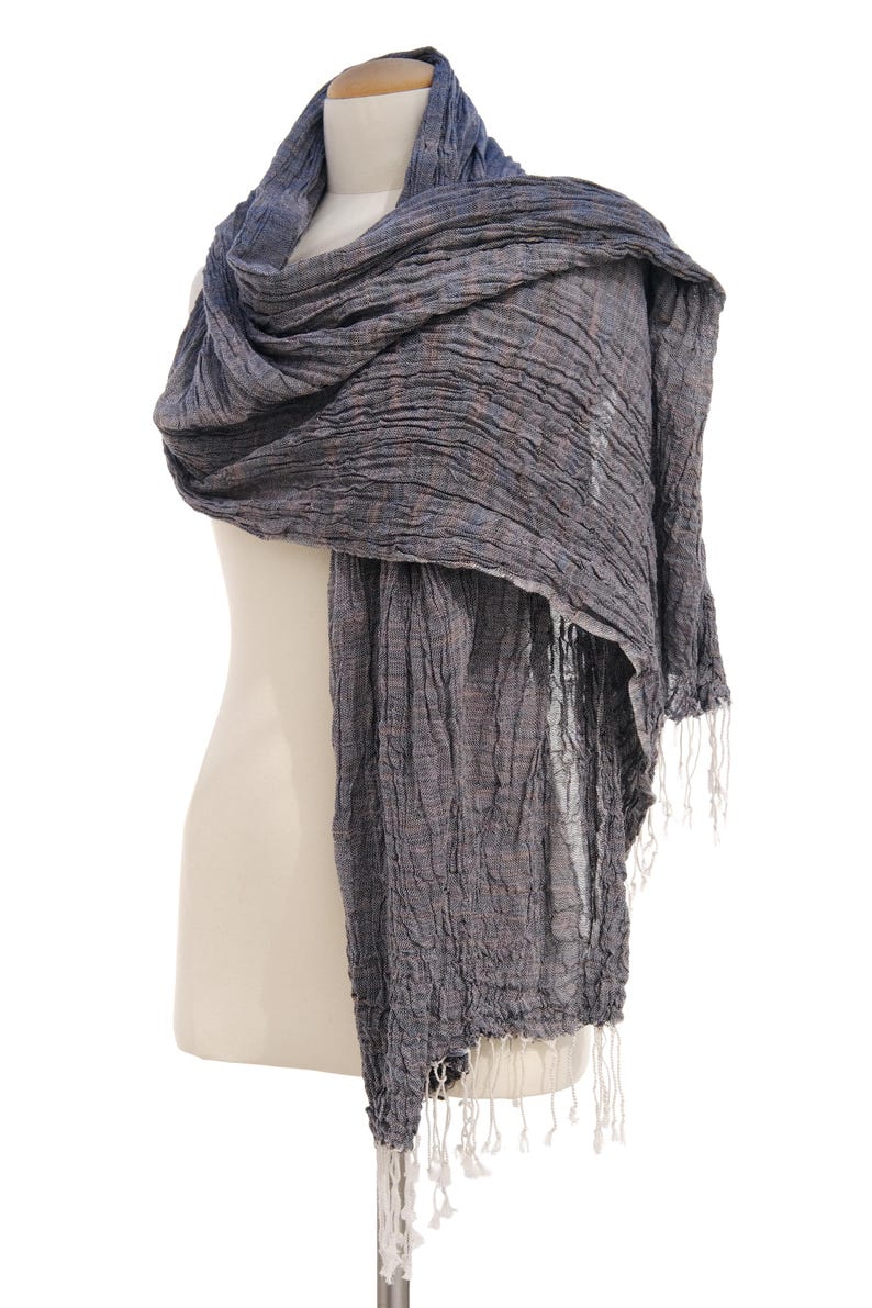May include: A draped, textured scarf in shades of gray and blue, displayed on a mannequin. The scarf has a crinkled appearance and is finished with white fringe at the ends. The fabric appears lightweight and suitable for layering.