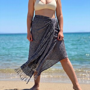 May include: A gray sarong with fringe detail is wrapped around the waist. The sarong is made of a textured fabric. The person is standing on a sandy beach with the ocean in the background. The person is wearing a beige bikini top.