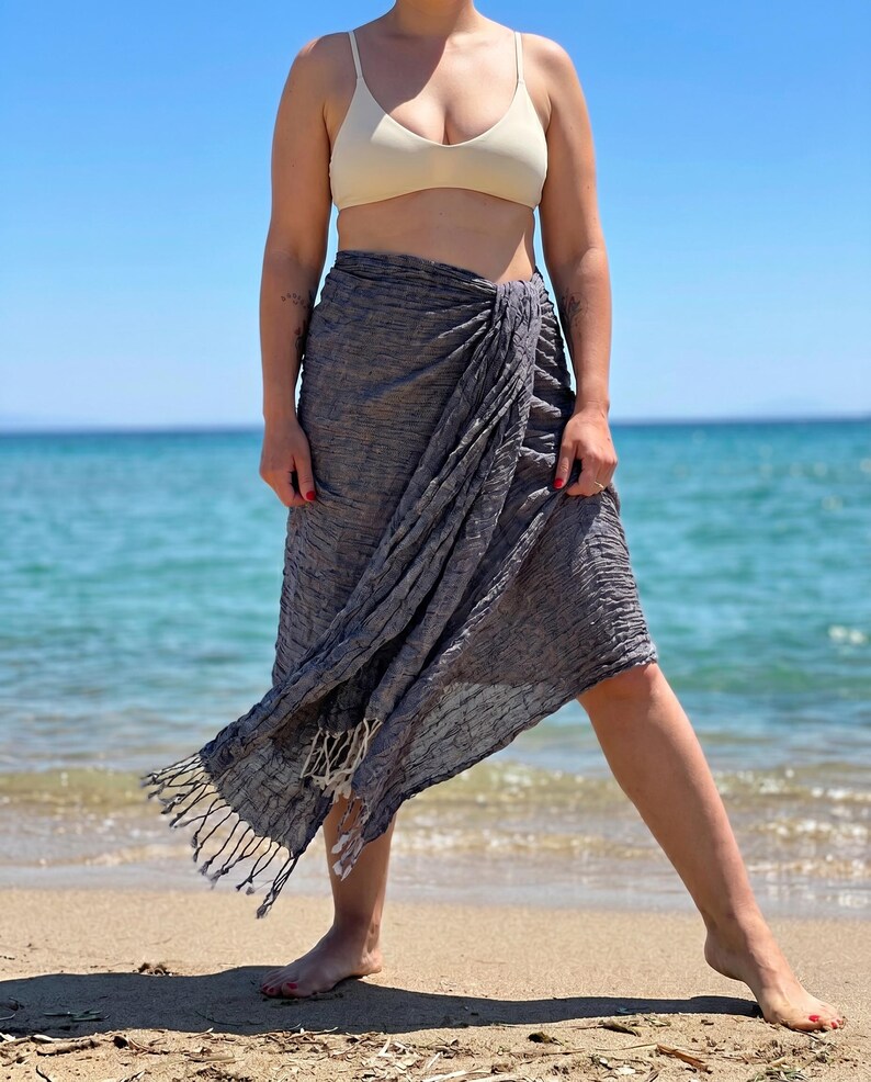 May include: A gray sarong with fringe detail is wrapped around the waist. The sarong is made of a textured fabric. The person is standing on a sandy beach with the ocean in the background. The person is wearing a beige bikini top.