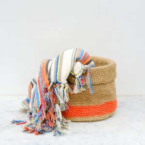 May include: A woven jute basket with a bright orange stripe and a colorful striped fabric with tassels inside.