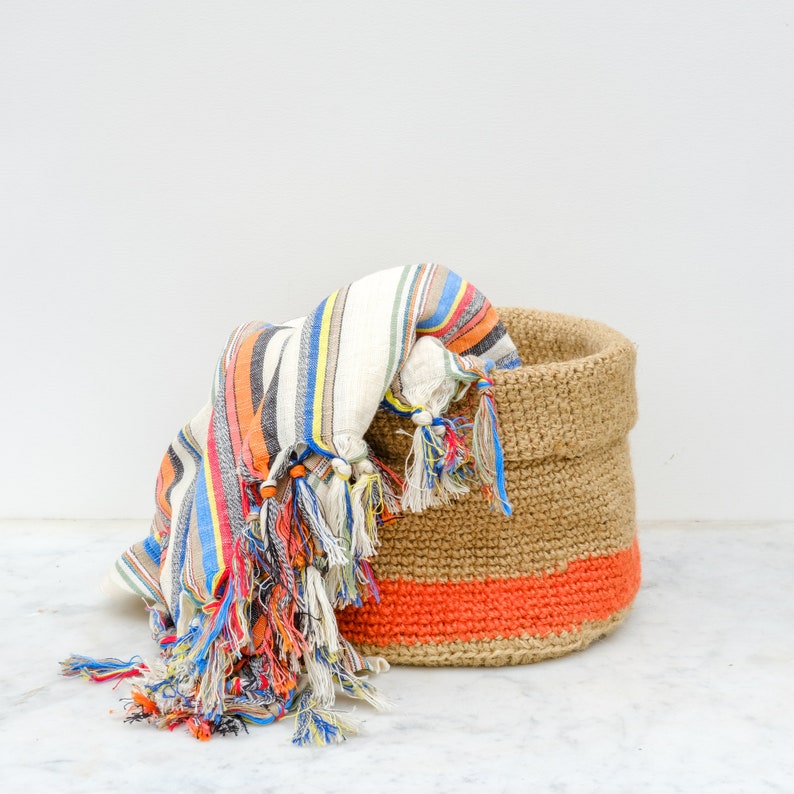 May include: A woven jute basket with a bright orange stripe and a colorful striped fabric with tassels inside.