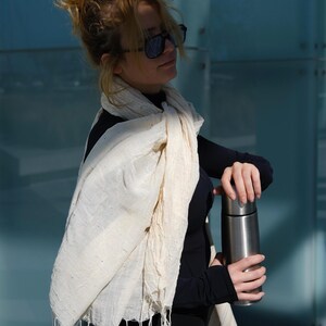 May include: A cream-colored scarf with fringe is draped over a person's shoulders. The scarf is paired with a black long-sleeved top and a stainless steel insulated water bottle. The background is a blue glass wall.