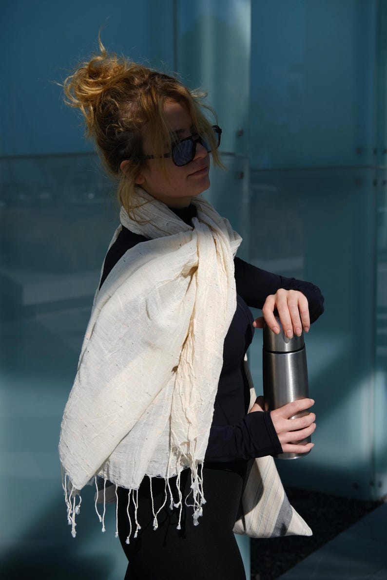 May include: A cream-colored scarf with fringe is draped over a person's shoulders. The scarf is paired with a black long-sleeved top and a stainless steel insulated water bottle. The background is a blue glass wall.
