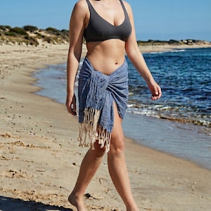May include: A woman wearing a dark gray bikini top and a blue sarong with cream-colored fringe walks on a sandy beach. The sarong is tied at the waist. The background shows the ocean and a clear blue sky.