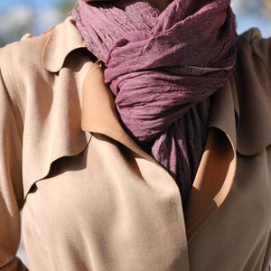 May include: A close-up of a tan trench coat with a mauve scarf. The scarf is wrapped around the neck and is a textured weave. The trench coat has a classic design with a notched collar and a double-breasted front.