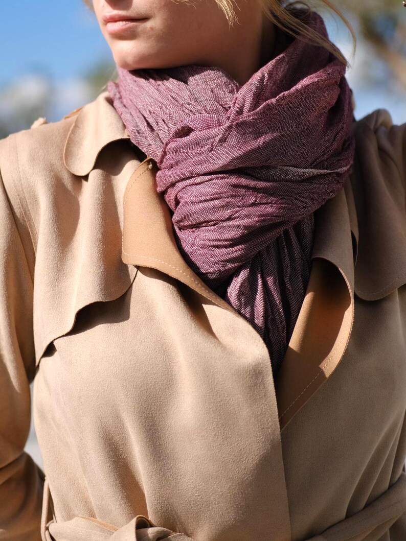 May include: A close-up of a tan trench coat with a mauve scarf. The scarf is wrapped around the neck and is a textured weave. The trench coat has a classic design with a notched collar and a double-breasted front.