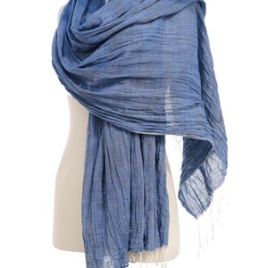 May include: A blue scarf with a textured appearance is draped over a mannequin. The scarf has a flowing design and is finished with white tassels at the ends. The scarf is made of a lightweight material.