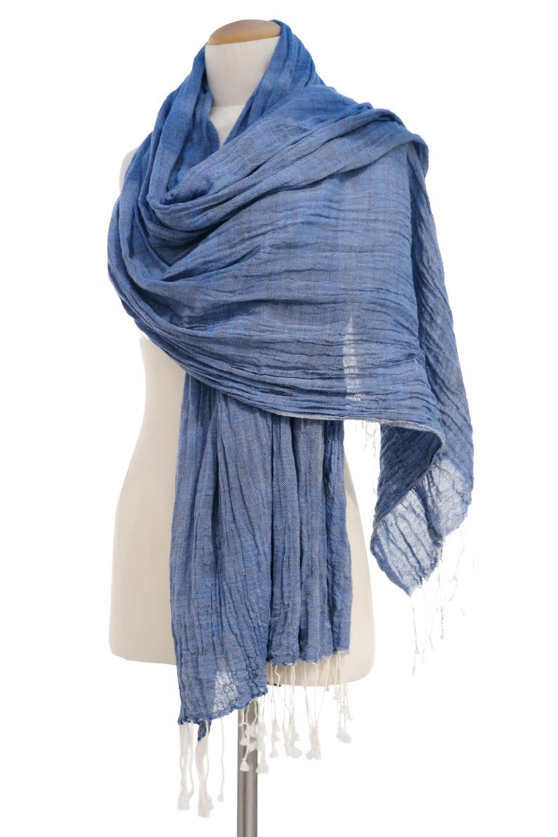 May include: A blue scarf with a textured appearance is draped over a mannequin. The scarf has a flowing design and is finished with white tassels at the ends. The scarf is made of a lightweight material.