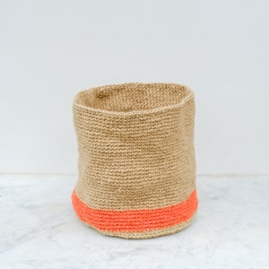 May include: A natural woven storage basket with a bright orange trim. The basket is made of a natural fiber and has a cylindrical shape.