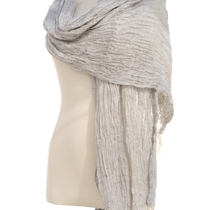 May include: A light gray scarf with a textured, crinkled appearance is draped over a mannequin. The scarf has a fringed edge and a subtle gradient of color, transitioning from a darker gray to a lighter shade.