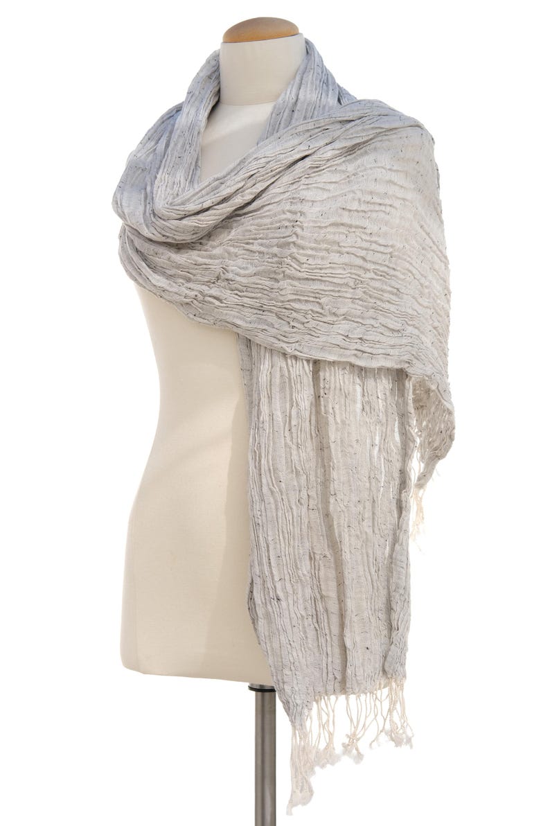 May include: A light gray scarf with a textured, crinkled appearance is draped over a mannequin. The scarf has a fringed edge and a subtle gradient of color, transitioning from a darker gray to a lighter shade.