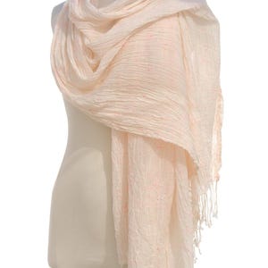 May include: A light peach-colored scarf draped over a mannequin. The scarf has a crinkled texture and is adorned with small, scattered pink accents. The ends of the scarf feature a fringe of white tassels, adding a delicate touch.