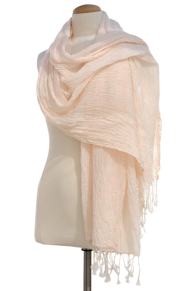 May include: A light peach-colored scarf draped over a mannequin. The scarf has a crinkled texture and is adorned with small, scattered pink accents. The ends of the scarf feature a fringe of white tassels, adding a delicate touch.