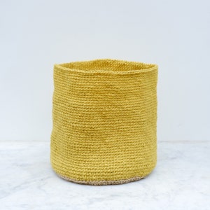 May include: A yellow crocheted storage basket with a natural colored trim. The basket is made of a woven material and has a cylindrical shape.