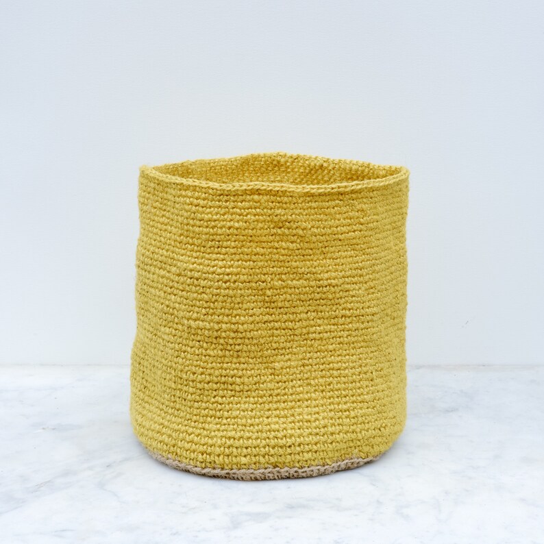 May include: A yellow crocheted storage basket with a natural colored trim. The basket is made of a woven material and has a cylindrical shape.