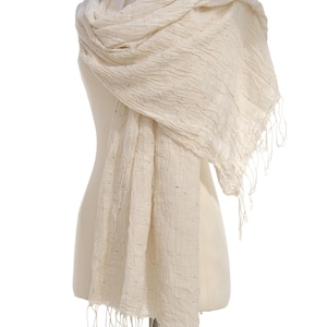 May include: Cream-colored scarf with a textured weave and fringe detail. The scarf is draped over a mannequin, showcasing its soft, flowing fabric. The neutral color and simple design make it a versatile accessory.