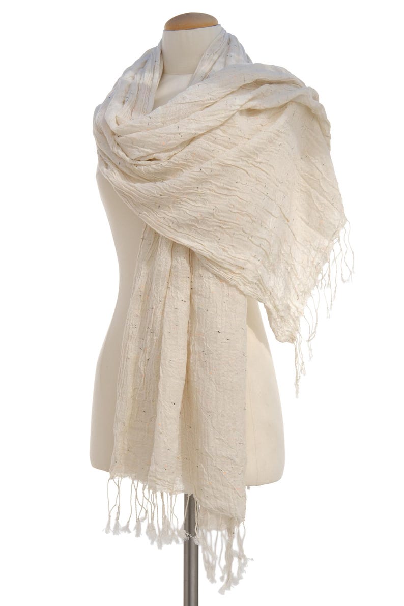 May include: Cream-colored scarf with a textured weave and fringe detail. The scarf is draped over a mannequin, showcasing its soft, flowing fabric. The neutral color and simple design make it a versatile accessory.