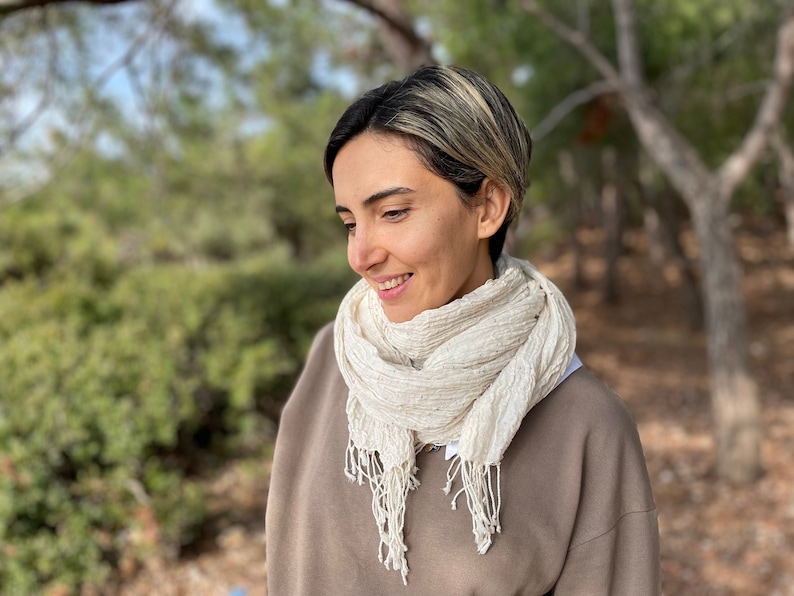 May include: A cream-colored scarf with a textured, crinkled appearance and fringed edges. The scarf is draped around the neck. The background is a blurred outdoor scene with trees and foliage.