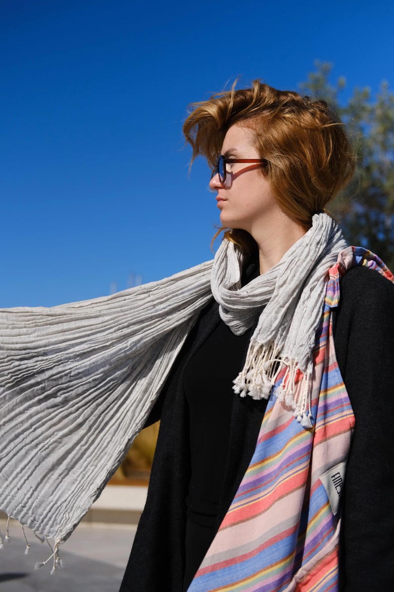 May include: A light gray, pleated scarf with white fringe is draped over a black top and coat. A striped tote bag with pink, blue, and orange hues hangs on the shoulder. The background is a clear blue sky.