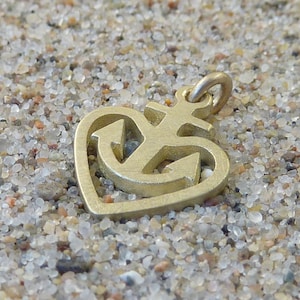 May include: A gold-coloured metal charm in the shape of a heart with an anchor inside. The charm is on a bed of small, white and brown pebbles.