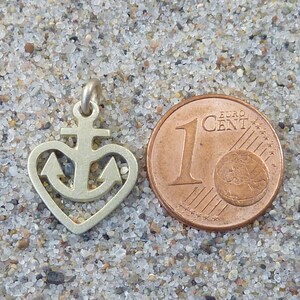 May include: A silver charm in the shape of a heart with an anchor inside. The charm is next to a 1 Euro Cent coin for size comparison.