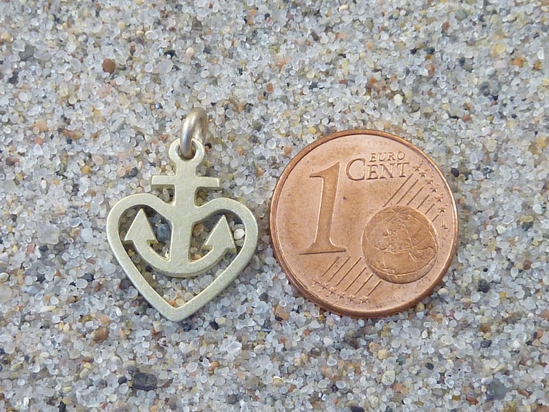 May include: A silver charm in the shape of a heart with an anchor inside. The charm is next to a 1 Euro Cent coin for size comparison.