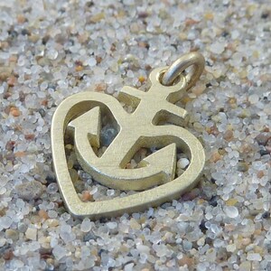 May include: A gold-coloured metal charm in the shape of a heart with an anchor inside. The anchor has a cross at the top.