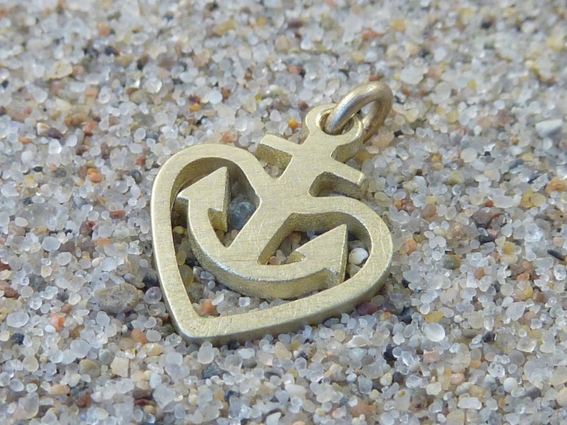May include: A gold-coloured metal charm in the shape of a heart with an anchor inside. The anchor has a cross at the top.