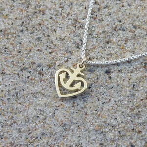 May include: A gold heart-shaped pendant necklace with an anchor design. The necklace is on a silver chain and is lying on a sandy surface.
