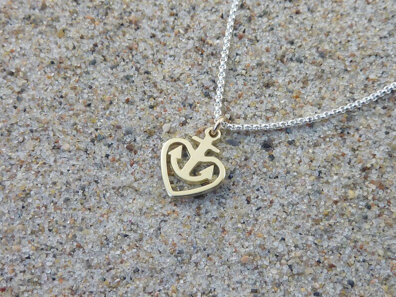 May include: A gold heart-shaped pendant necklace with an anchor design. The necklace is on a silver chain and is lying on a sandy surface.