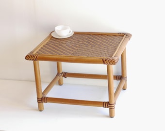 May include: A square, light brown rattan side table with a woven top. A white teacup and saucer sit on the table. The table is approximately 46 cm tall and 46 cm wide.
