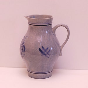 Könnte beinhalten: Ein grauer Keramik Krug mit blau-weißem Design. Der Krug hat einen runden Körper und einen Henkel.