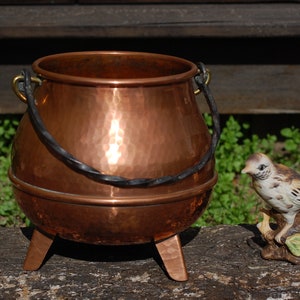 Könnte beinhalten: Ein Kupferkessel mit einem schwarzen Henkel und drei Beinen steht auf einer Holzoberfläche neben einer Porzellan-Vogelfigur.