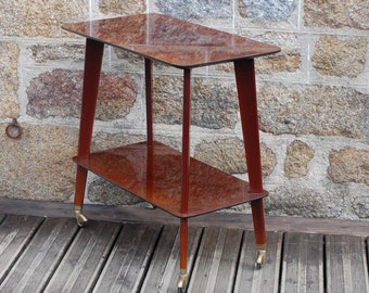 May include: A two-tiered mahogany wood serving cart with gold wheels. The cart has a rectangular top and bottom shelf and is supported by four angled legs.