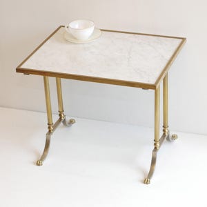 May include: A small side table with a white marble top and a brass frame. The table has four brass legs, each with a decorative foot. A white teacup and saucer sit on the table's surface. The table is approximately 60 cm in height.