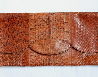 May include: A rectangular, brown clutch with a snakeskin pattern. The clutch has a flap closure and a decorative oval accent in the center. The overall design is a mix of textures and shades of brown.