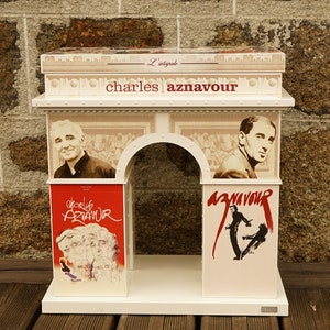 Op de afbeelding: Een wit model van de Arc de Triomphe in Parijs, Frankrijk, met een rood, wit en zwart ontwerp met de naam "Charles Aznavour" en afbeeldingen van de zanger.