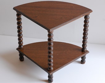 May include: A two-tiered corner table with a dark brown wood finish. The table has a rounded, semi-circular top and bottom shelf. The legs are made of turned wood with a beaded design.