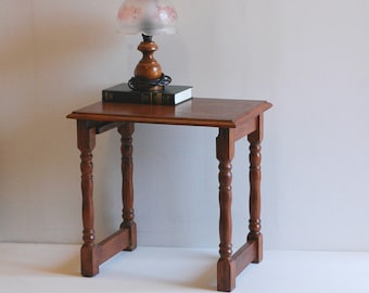 May include: A small, antique wooden side table with turned legs. A vintage lamp with a floral patterned shade and a book rest on the table's surface. The table is a warm brown color.